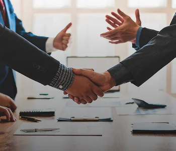 Two individuals in business attire shaking hands over a conference table during a meeting.