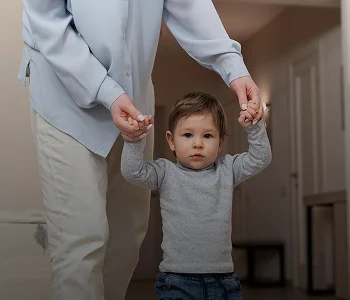 Adult holding hands of a toddler wearing gray sweater and blue jeans indoors.