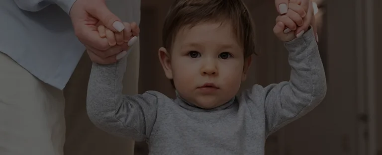 Toddler wearing a gray sweater holding hands with an adult while learning to walk indoors.