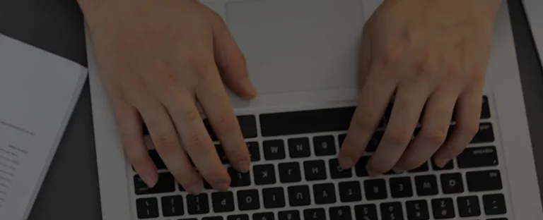 Close-up of hands typing on a black and silver laptop keyboard beside an open book.