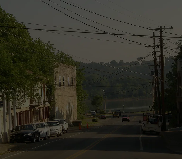 Quiet street with parked cars, utility poles, and a wooded hillside in the background.