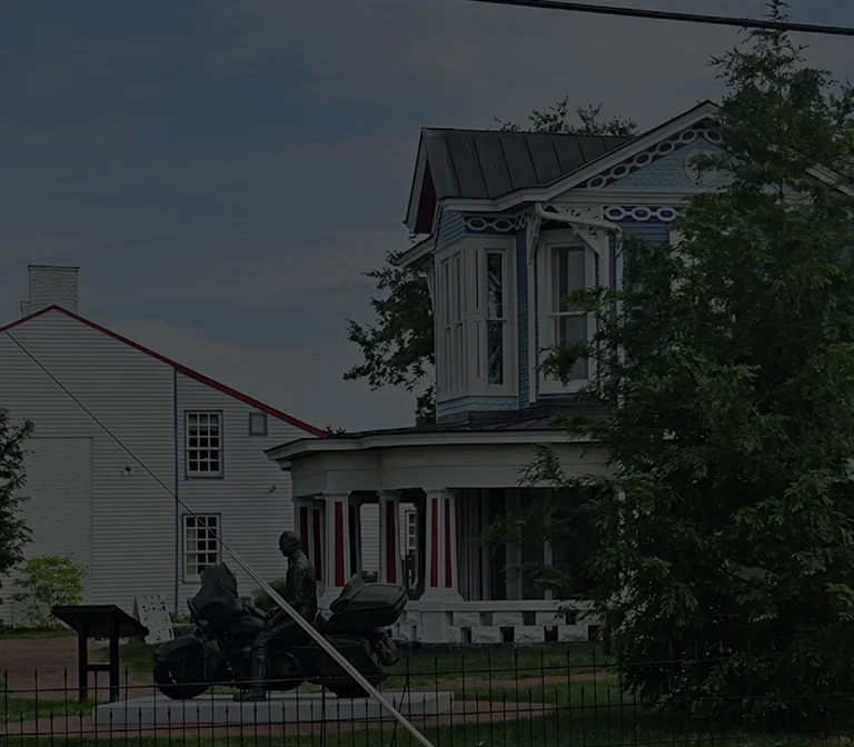 Two-story blue Victorian house with white trim partially obscured by trees, adjacent white building, and black motorcycle statue in front.