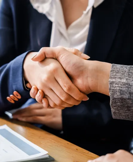 Two professionals in business attire shaking hands over a wooden table with documents.