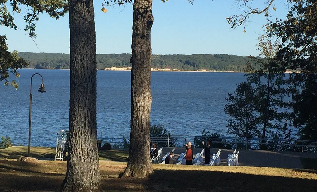 Scenic lakeside view with large trees, blue water, distant forest, and people near white chairs.