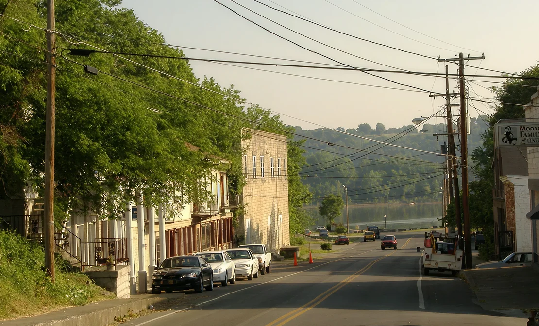 Quiet small-town street with parked cars, trees, utility poles, and distant hills.
