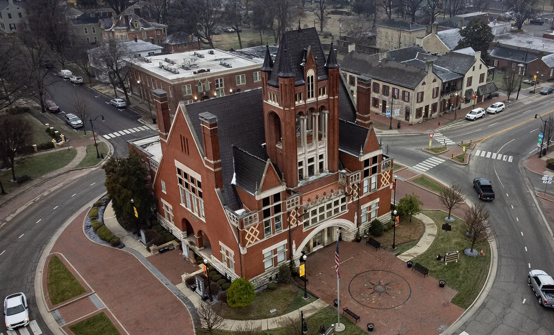 Aerial view of a large red-brick historic building with pointed towers surrounded by roads and sidewalks.