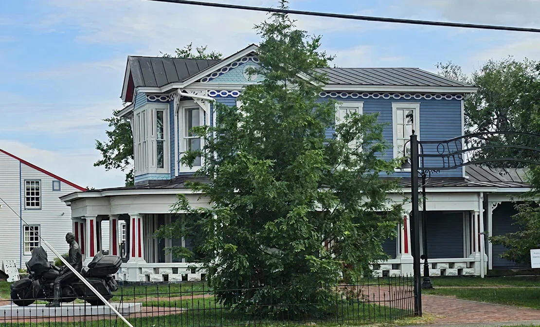 Historic two-story blue house with white trim, surrounded by trees and a black iron fence.