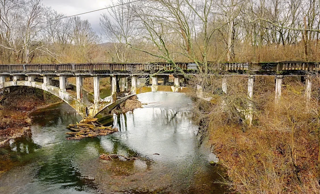 A weathered concrete bridge spans a calm river surrounded by leafless trees in late autumn.