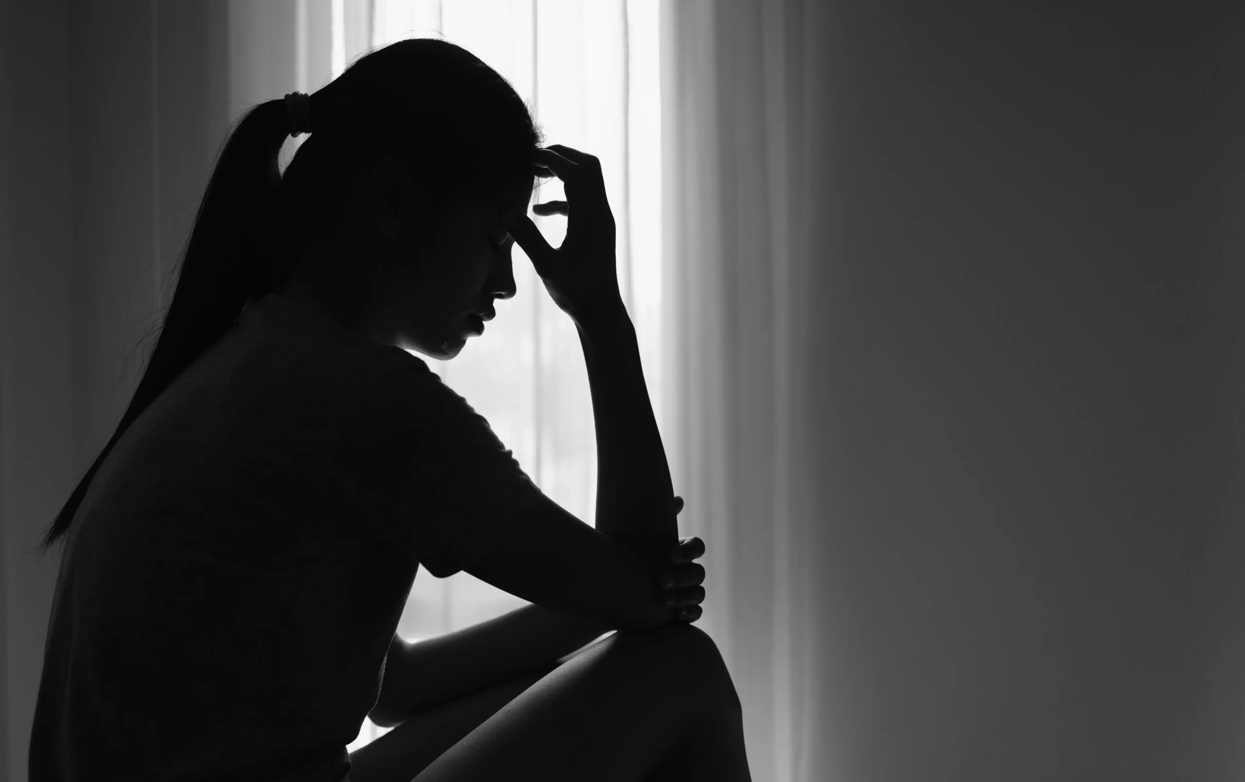 Silhouetted woman with ponytail sitting and holding her forehead, backlit by a sheer curtain.