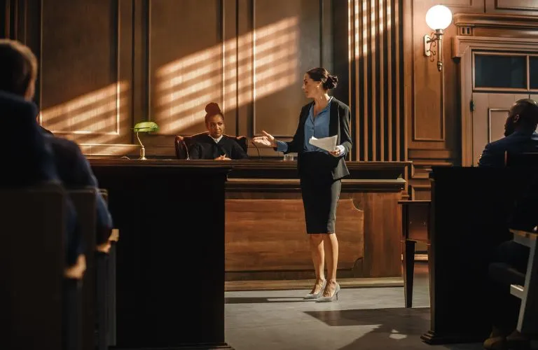Female lawyer presenting a case in a wood-paneled courtroom with a judge and audience.