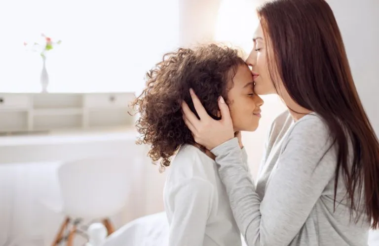 Woman with long brown hair gently kisses the forehead of a curly-haired child in a bright room.