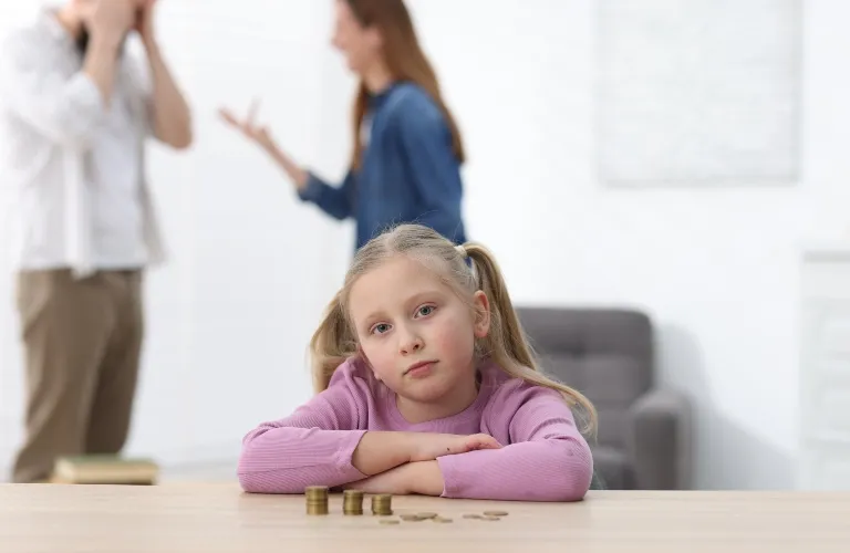 Young girl in purple sweater resting on table with coins, parents arguing blurred behind.