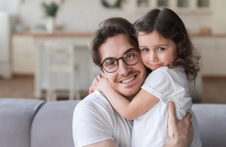 Man with glasses and white shirt hugging young girl with curly brown hair and white shirt on gray couch.