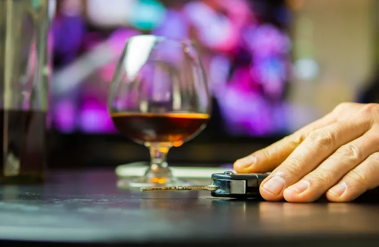 Close-up of a hand reaching for car keys on a table with a glass of amber-colored liquor in the background.