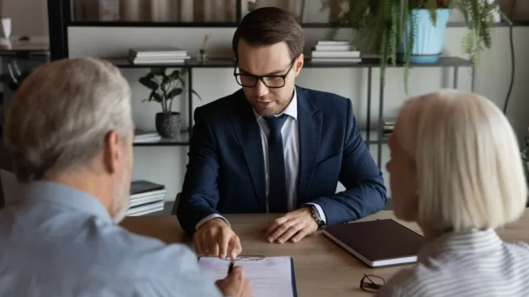 Young professional man in navy suit and glasses explaining documents to elderly couple at office desk.