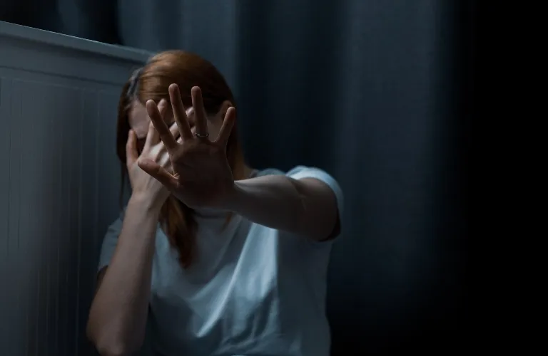 Person with red hair in white shirt covering face with one hand and extending other hand forward in dark room.