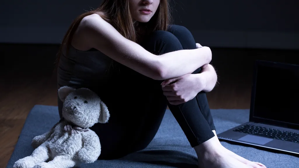 Young woman sitting on a mat hugging her knees, with a white teddy bear and a laptop nearby.