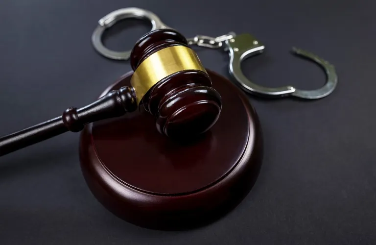 Wooden judge's gavel with gold band resting on sound block beside metal handcuffs on black surface.