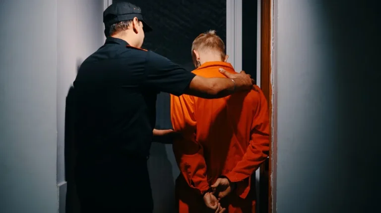A security officer escorting a person in an orange jumpsuit with hands cuffed behind back.