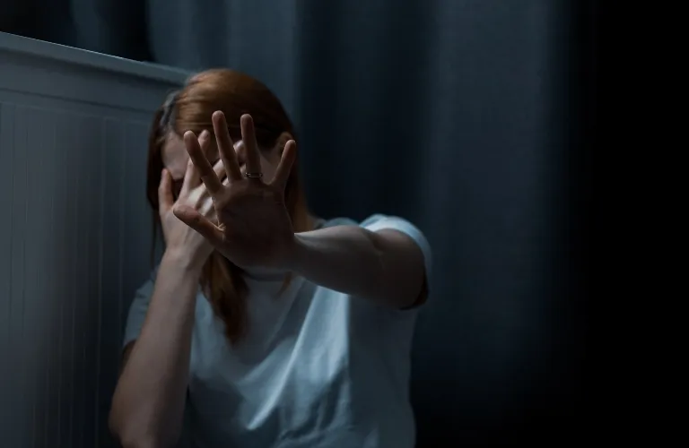 Woman with red hair in white shirt shielding face with hand in dimly lit room.