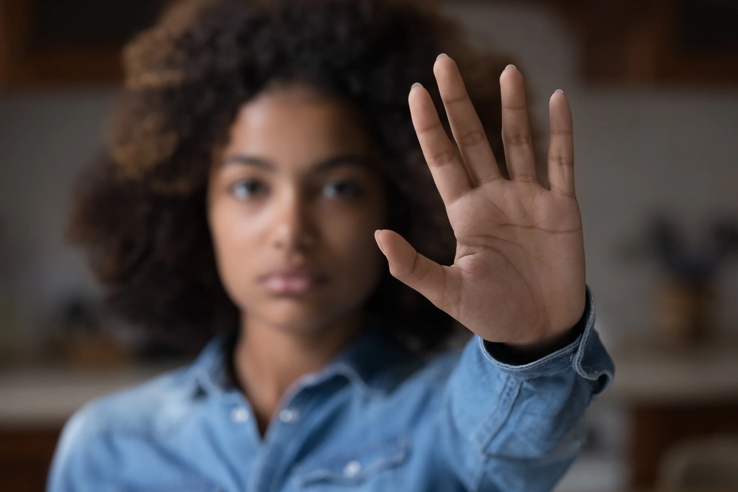 Young woman with curly hair wearing a blue shirt, holding hand up towards camera.