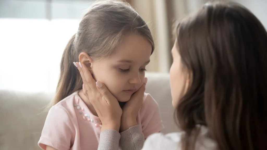 Woman gently holding a young girl's face, comforting her indoors with soft natural light.